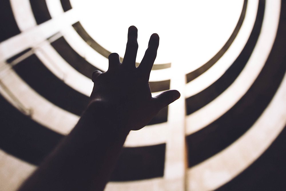 Una mano se eleva hacia una luz brillante, enmarcada por balcones circulares concéntricos o pisos superiores. La imagen está tomada desde un ángulo bajo, creando una sensación de aspiración o búsqueda de la luz.