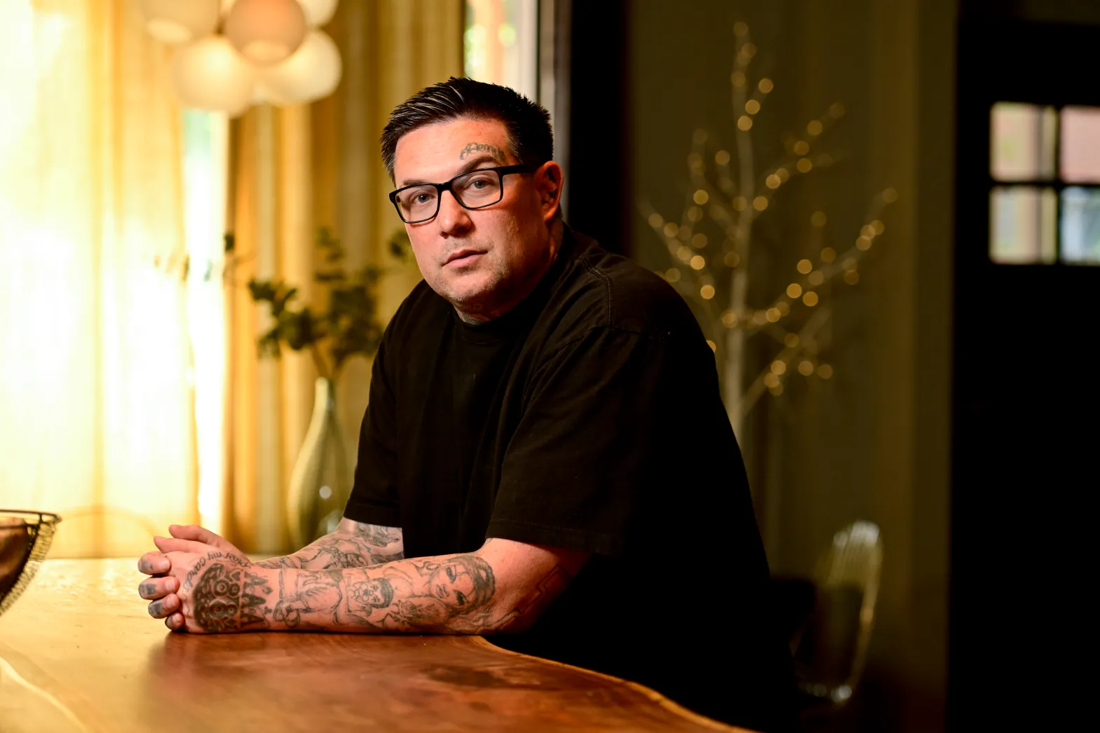 Una persona con el pelo corto y oscuro, gafas y múltiples tatuajes en los brazos está apoyada en una mesa de madera. Lleva una camiseta negra con el lema "CU Boulder". La habitación, suavemente iluminada con una lámpara encendida y adornos, crea una atmósfera acogedora, perfecta para los debates de divulgación comunitaria.
