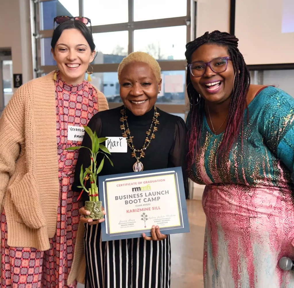Three people smile at the camera. The person in the middle holds a certificate and a small plant. They are in a room with large windows. The group appears cheerful and is dressed in colorful attire.
