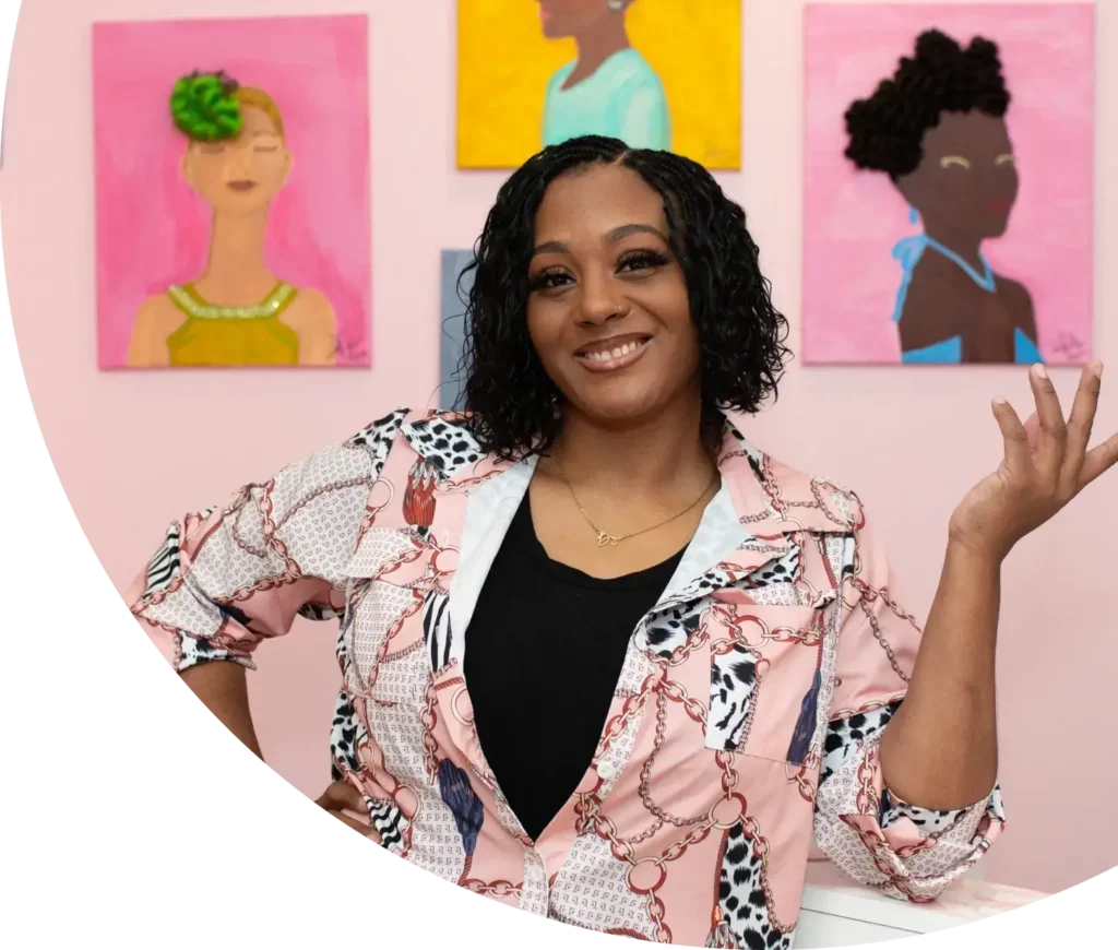 A woman smiles confidently in front of colorful paintings featuring different women. She wears a patterned pink jacket over a black top, has curly hair, and gestures with her left hand. The background is a pink wall displaying the vibrant artwork.
