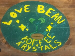 A hand-painted sign on wood features "Love Bean Koffee Krystals" in vibrant yellow. A sprouting coffee bean takes center stage, enveloped by star-like accents that complete the charming design.