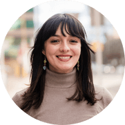 Smiling person with long dark hair wearing a turtleneck sweater and dangling earrings, standing outdoors with a blurred urban background.