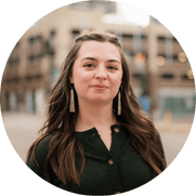 Young woman with long brown hair and tassel earrings, smiling slightly, stands outdoors in a blurred urban setting. She is wearing a dark green top, and buildings are visible in the background.