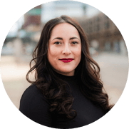 A person with long dark hair and red lipstick is wearing a black top. They are standing outdoors with a blurred cityscape in the background.