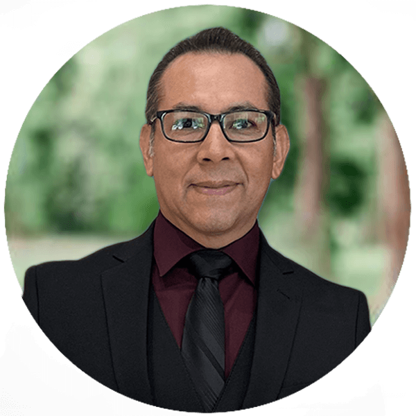 A person wearing glasses, a black suit, and a burgundy shirt with a black tie is smiling. The background is a blurred outdoor scene with green foliage.