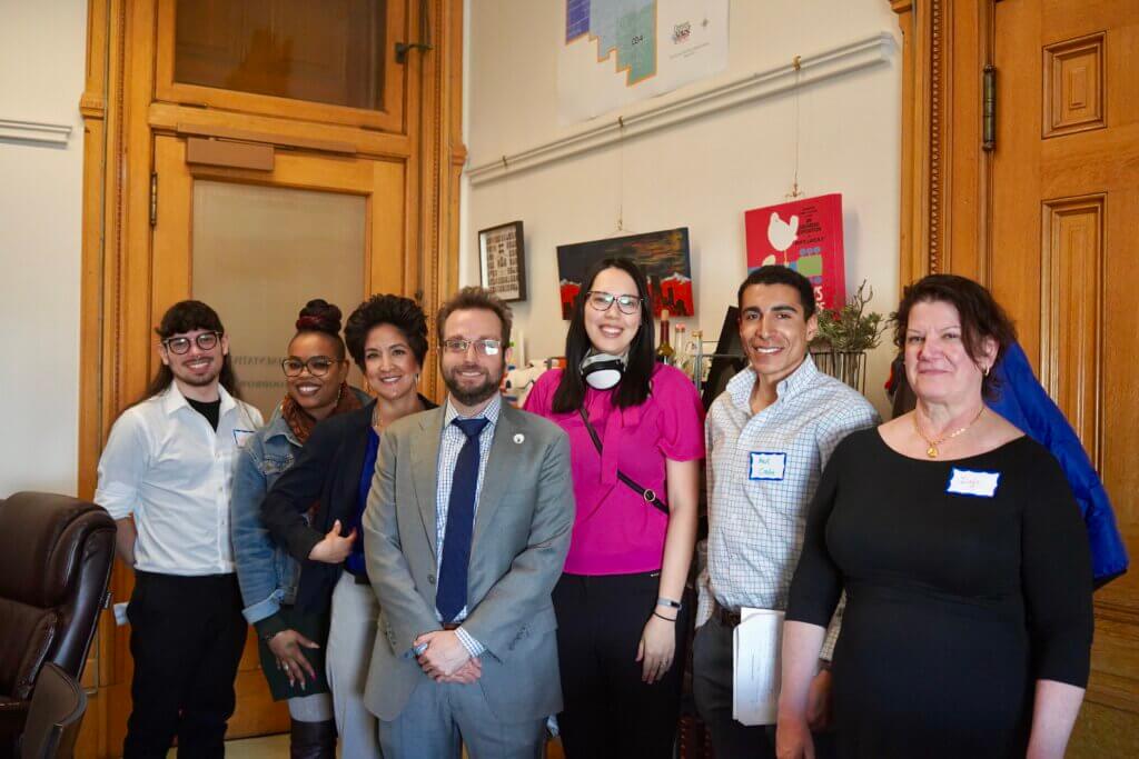 A diverse group of seven entrepreneurs, both men and women, are standing indoors, smiling at the camera. They are dressed in business casual attire and are in an office setting with artwork and wooden doors in the background, capturing the spirit of Entrepreneur Day perfectly.
