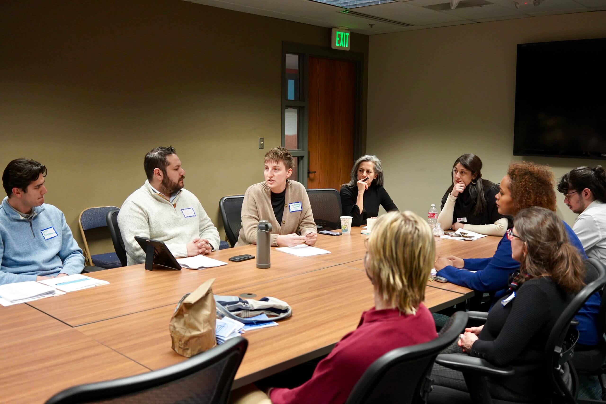 A diverse group of entrepreneurs sits around a large table in a meeting room, engaging in lively discussion.