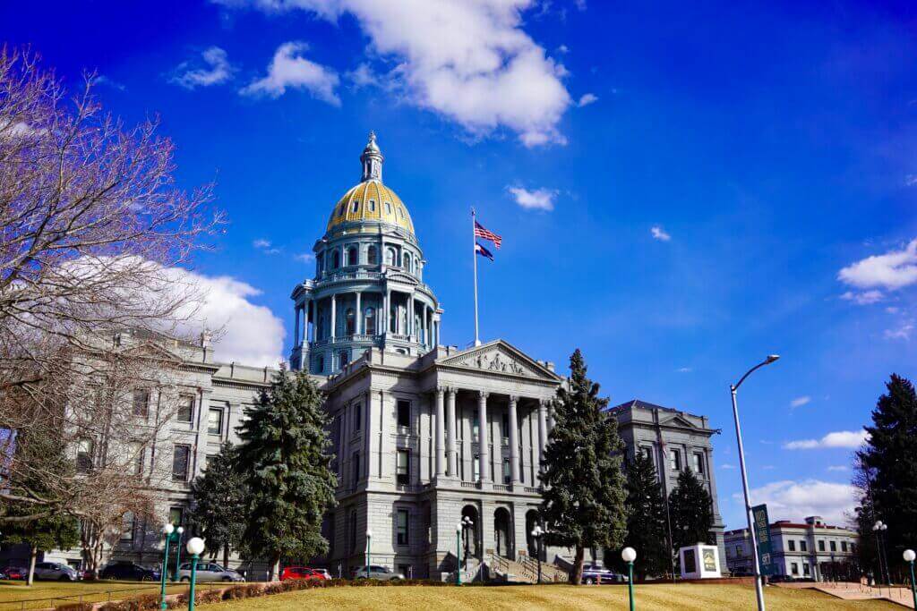 The image shows a historic building with a prominent golden dome and a tall flagpole flying the American flag, echoing the spirit of Entrepreneur Day of Action. The structure is surrounded by trees and set against a vivid blue sky with scattered clouds, making voices heard in this vibrant setting.