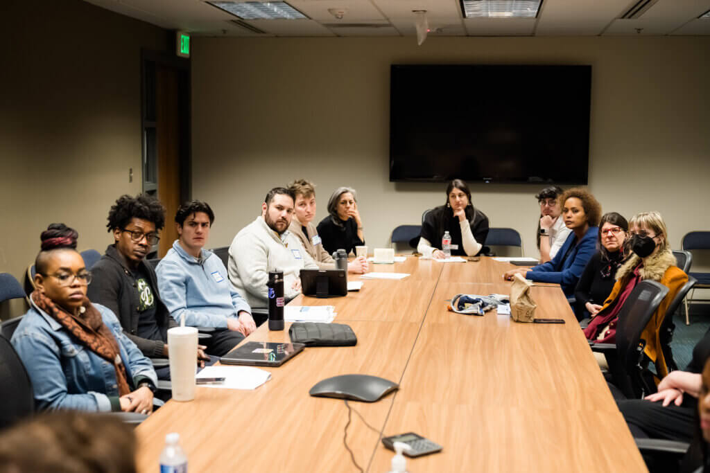 A group of entrepreneurs is seated around a long conference table in a meeting room, actively engaged in a discussion. Notebooks and drinks are scattered on the table, and a large screen is mounted on the wall, reminiscent of an Entrepreneur Day event.