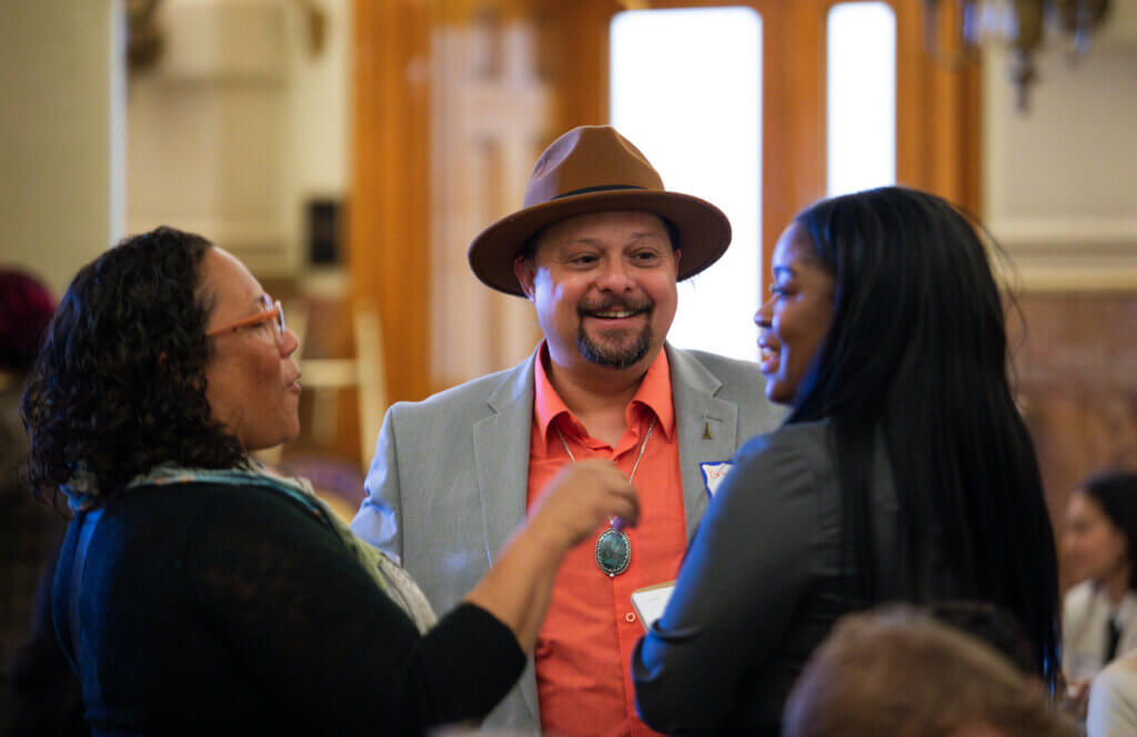Three individuals stand in conversation, their voices clearly heard. A person in a hat and light jacket smiles warmly while speaking to the other two. One has curly hair, glasses, and gestures animatedly; the other with long hair wears a dark jacket. The scene unfolds indoors during Entrepreneur Day of Action.