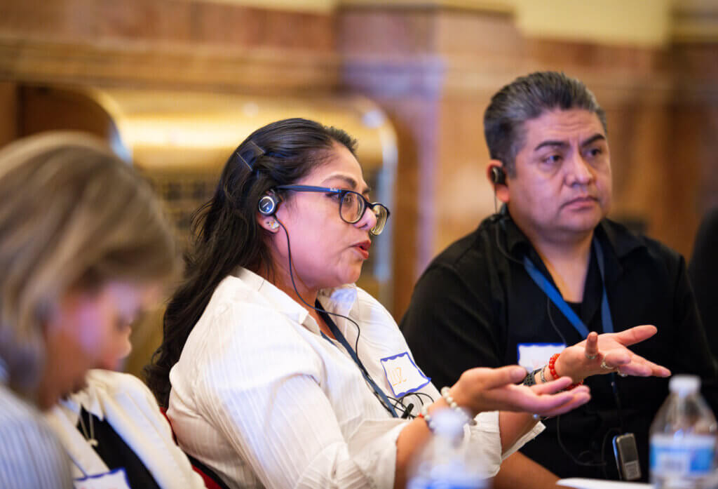 A woman wearing a headset speaks passionately, gesturing as she sits at a table with entrepreneurs. A man beside her listens attentively, embodying the spirit of an Entrepreneur Day of Action. The setting is indoors, suggesting a dynamic meeting or seminar where voices are heard and shared.