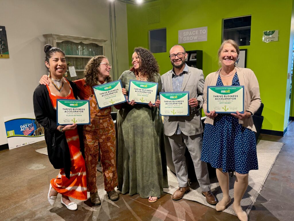 Five smiling people stand indoors holding certificates that read “Thrive Business Accelerator.” They are dressed in colorful, professional clothing and stand in front of a green and beige wall with plants and a "Space to Grow" sign.
