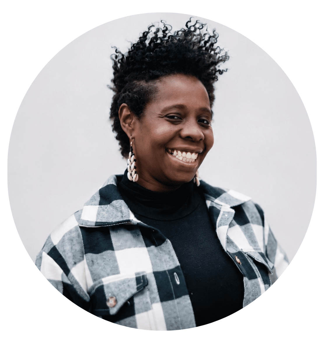 A person with short curly hair smiles, wearing earrings, a black turtleneck, and a black-and-white plaid jacket, standing against a light gray background—perfect for an About RMMFI feature.
