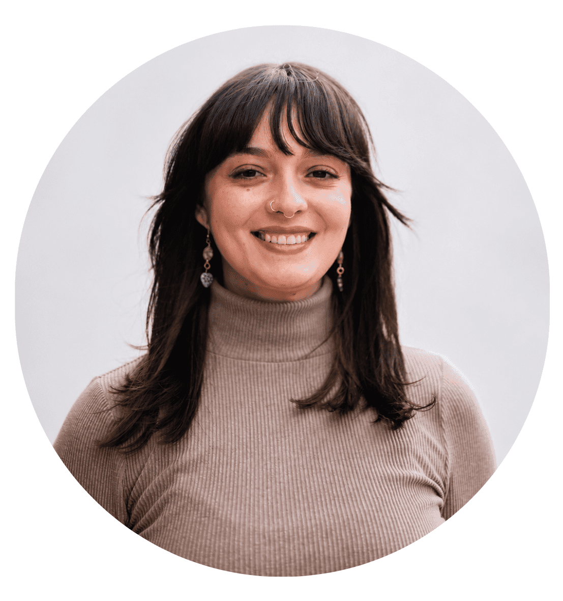 A woman with medium-length dark hair and bangs is smiling. She is wearing a beige turtleneck sweater and green earrings. Framed in a circular crop, the light, neutral background gives this About RMMFI photo a warm and approachable feel.