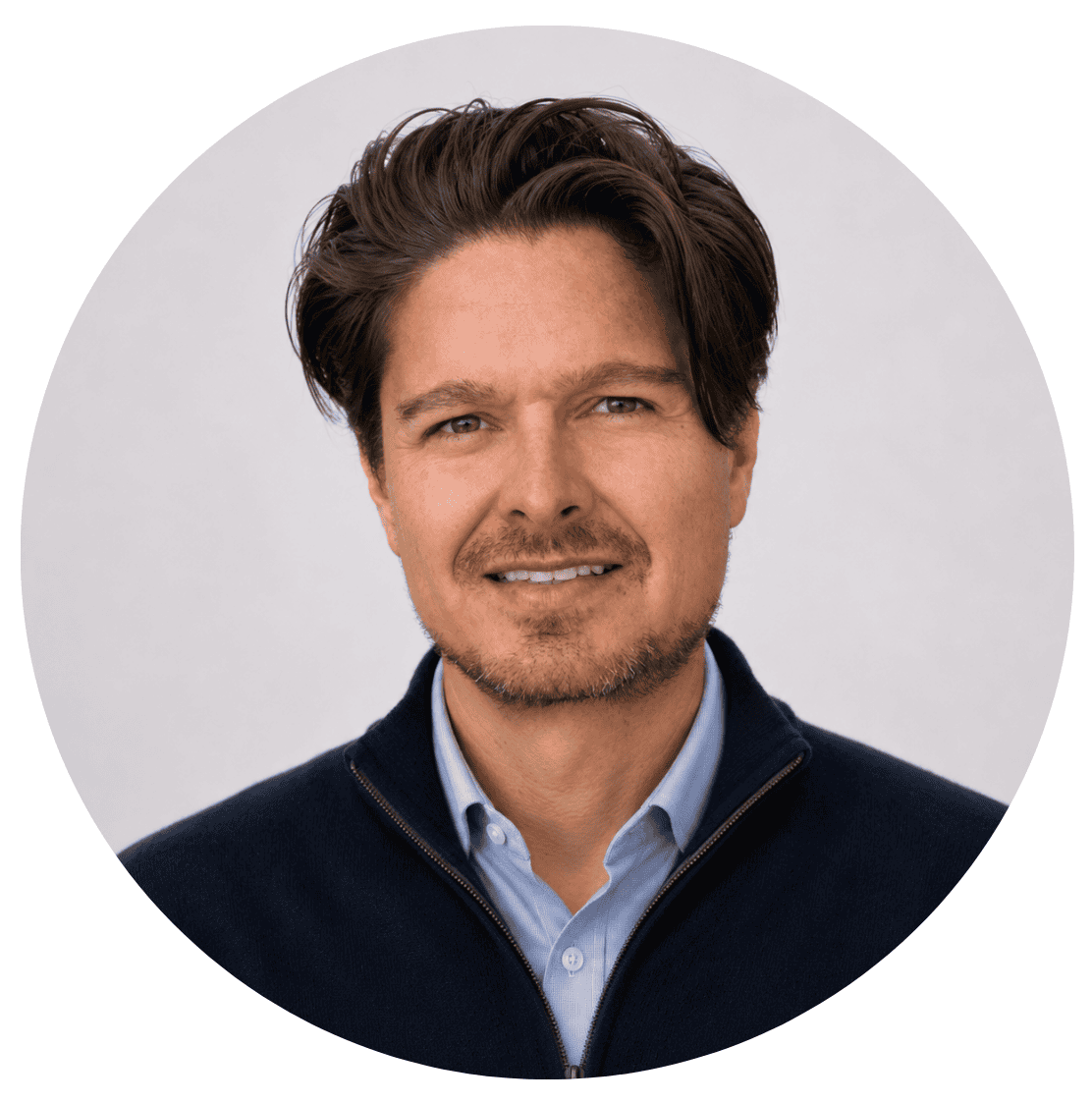 A man with medium-length brown hair, light facial hair, and a slight smile is wearing a navy jacket over a light blue shirt, posing in front of a plain, light background—perfectly representing the approachable spirit found at About RMMFI.